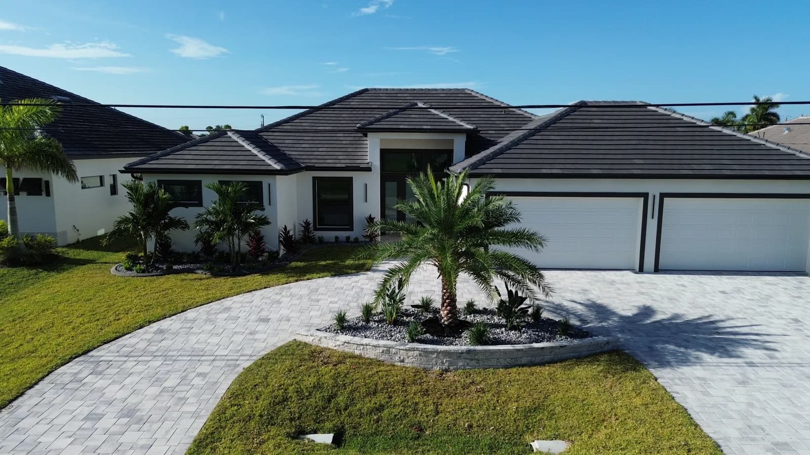 Front of a new white stucco home in Southwest Florida with a curved paver driveway, a stacked-stone planter with a palm at its center and fresh entry beds, installed by Blue Collar Q
